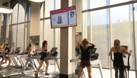 Students on treadmills in a campus gym with a branded ad displayed on the overhead TV screen